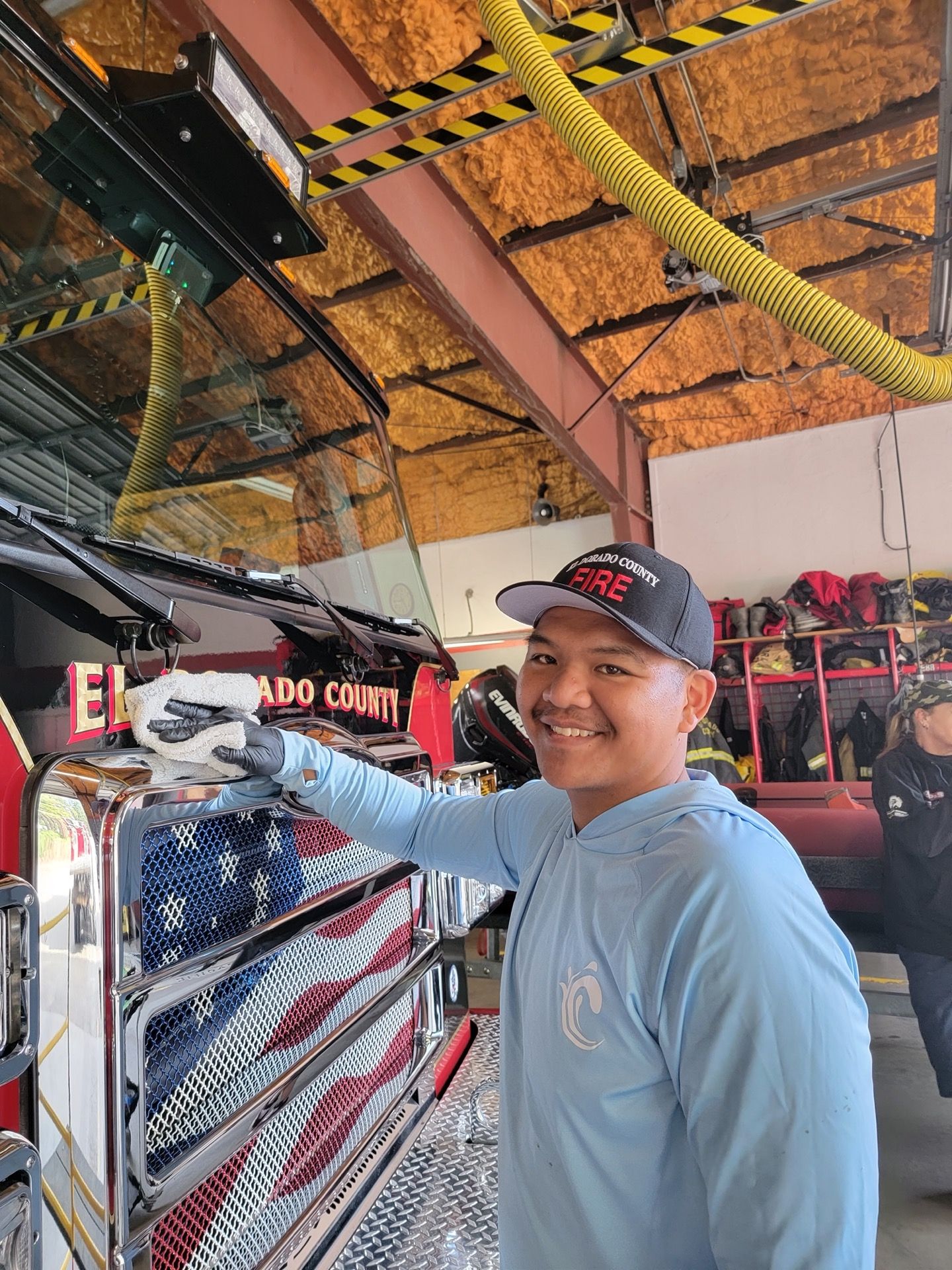 Firefighter, smiling, cleaning fire truck with American flag design. Outdoors, garage setting.