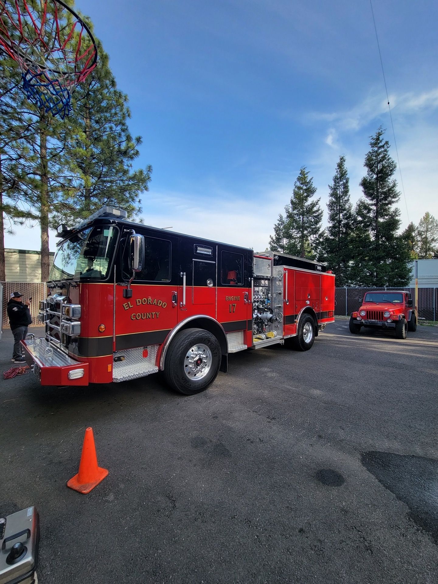 Red fire truck with black accents parked outside under a blue sky, next to a red Jeep.