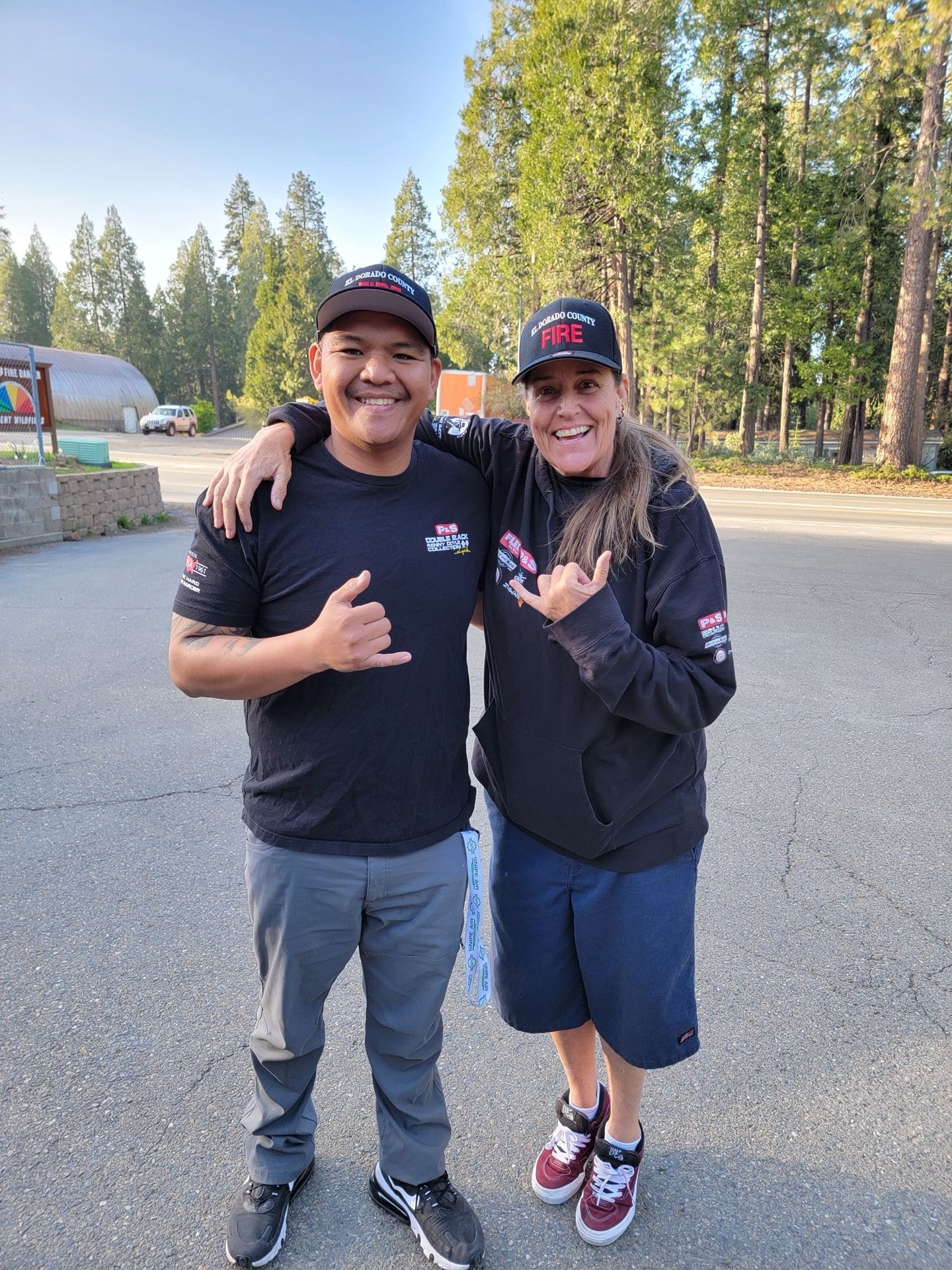 Two people smiling, embracing, outdoors. Both wearing black hats and shirts, gray pants, and shorts.