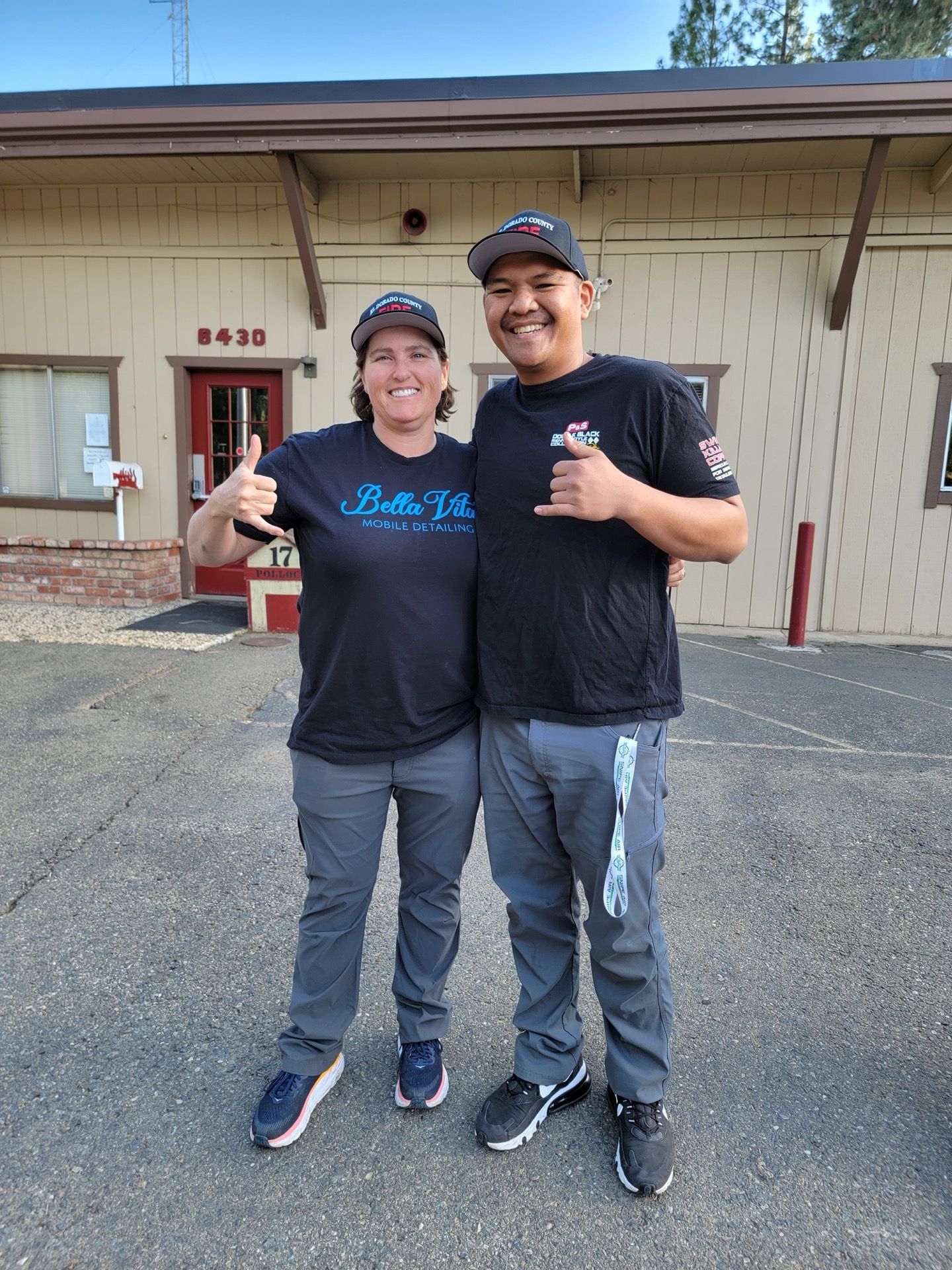 Two people in matching hats and attire smile and give a thumbs up/hang loose sign outside a building.