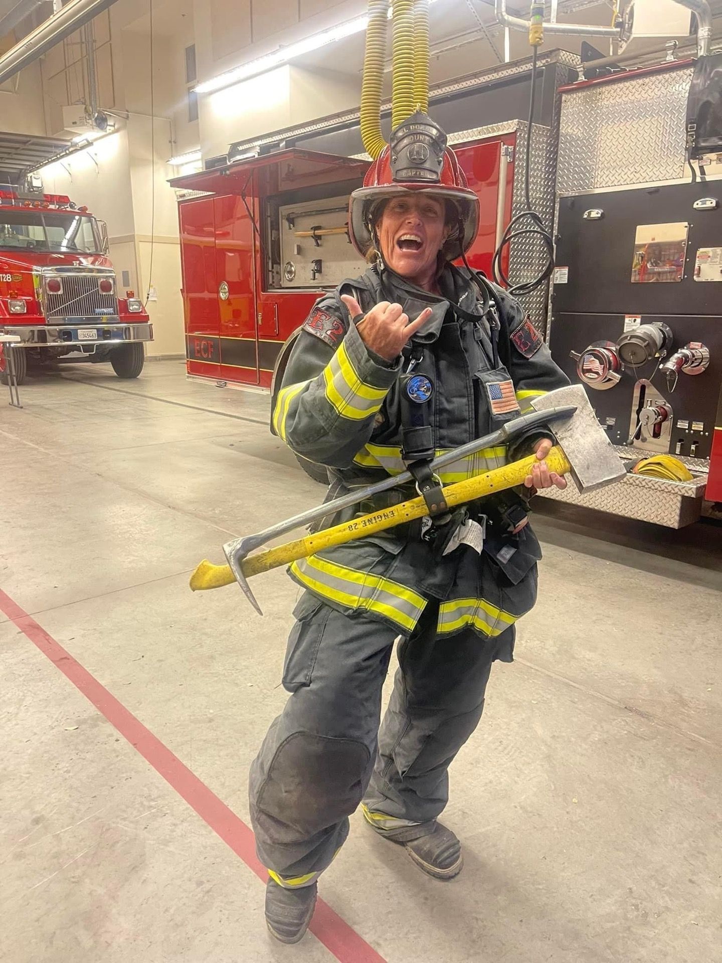 Firefighter in turnout gear holding an axe, giving the shaka sign, inside a fire station.