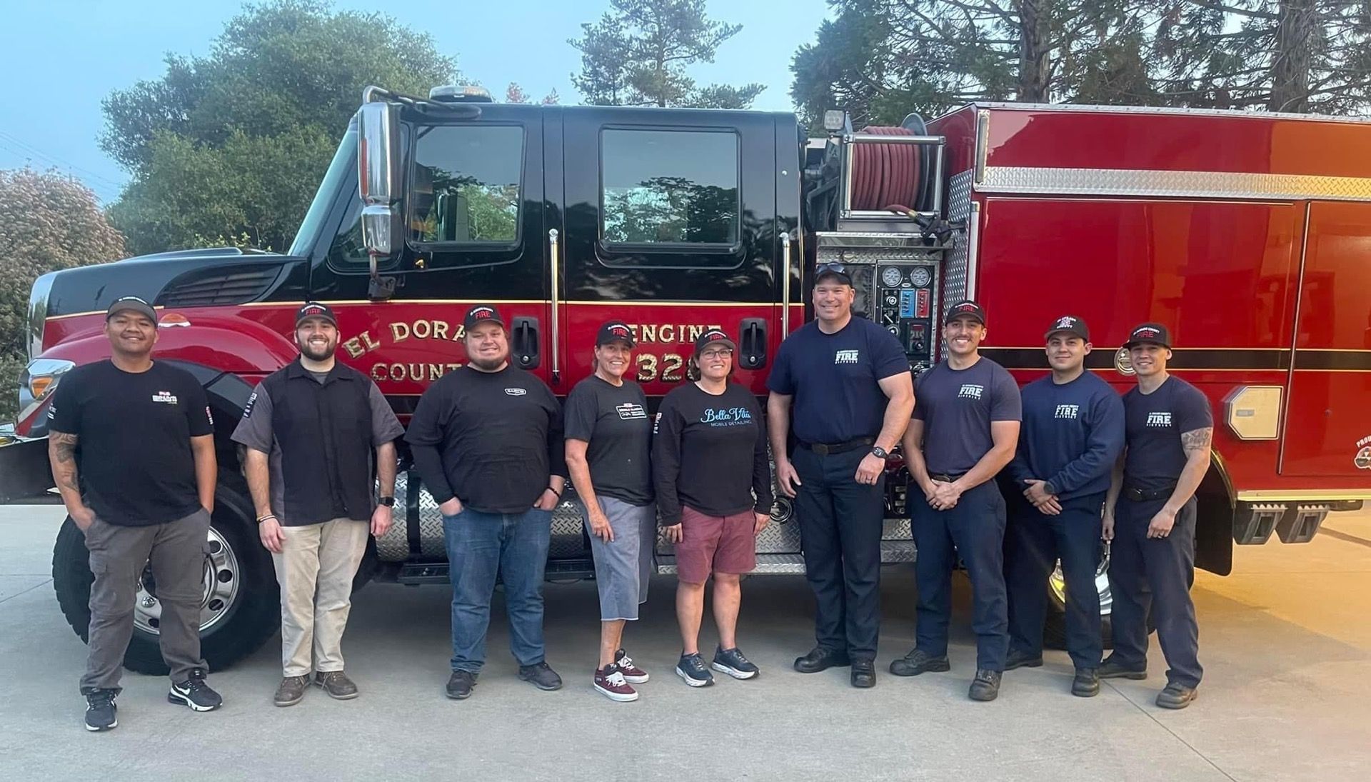 Firefighters posing with a red fire truck in front of a building; outdoor shot, evening.