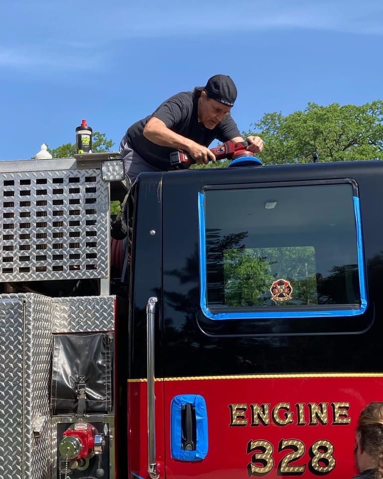 Man polishes a fire truck's black roof under a blue sky.