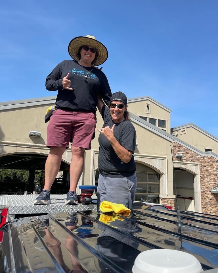 Two people on a truck roof, smiling, giving hang loose signs. Sunlit day. Building background.