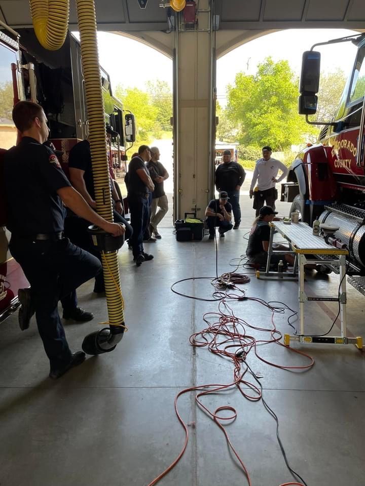Firefighters working in a fire station bay with a fire truck, some observing, tools and cords visible.