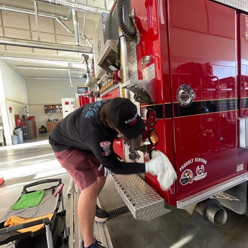 Person cleaning a red fire truck, wearing a black hat and shirt, in a garage.