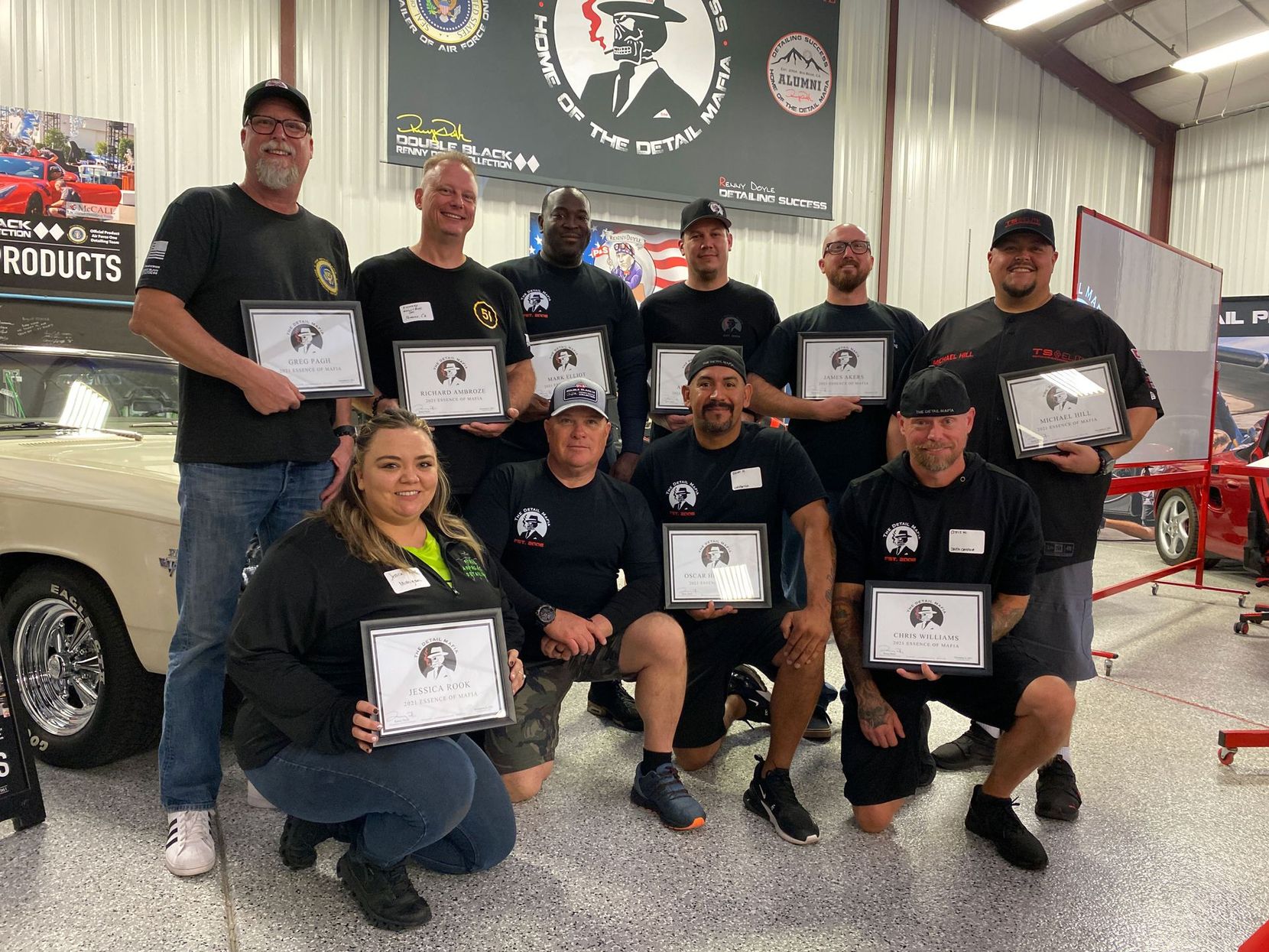 Group of people, in a workshop setting, hold framed certificates; smiling.