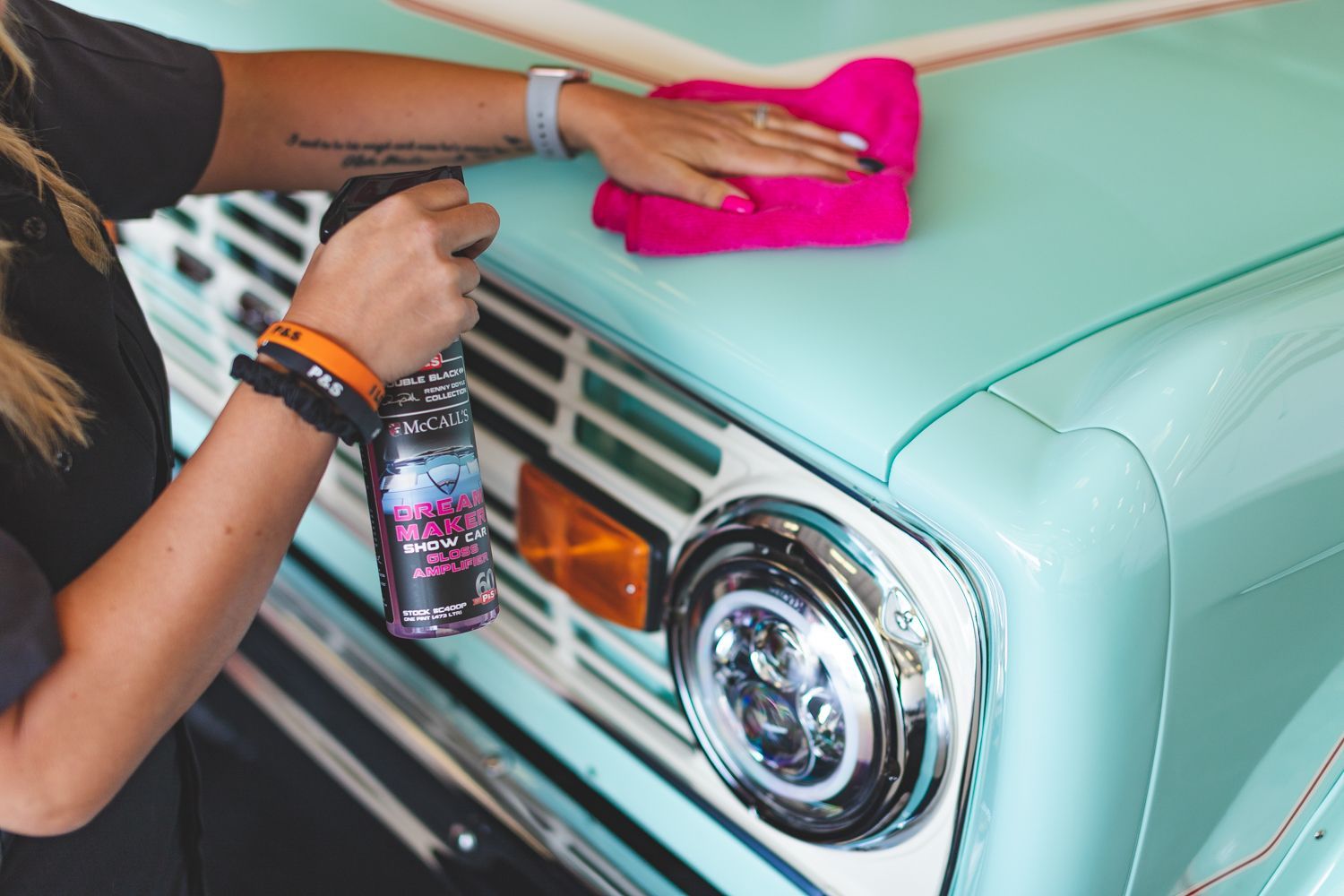 Person spraying cleaner on a turquoise car hood, wiping it with a pink cloth.
