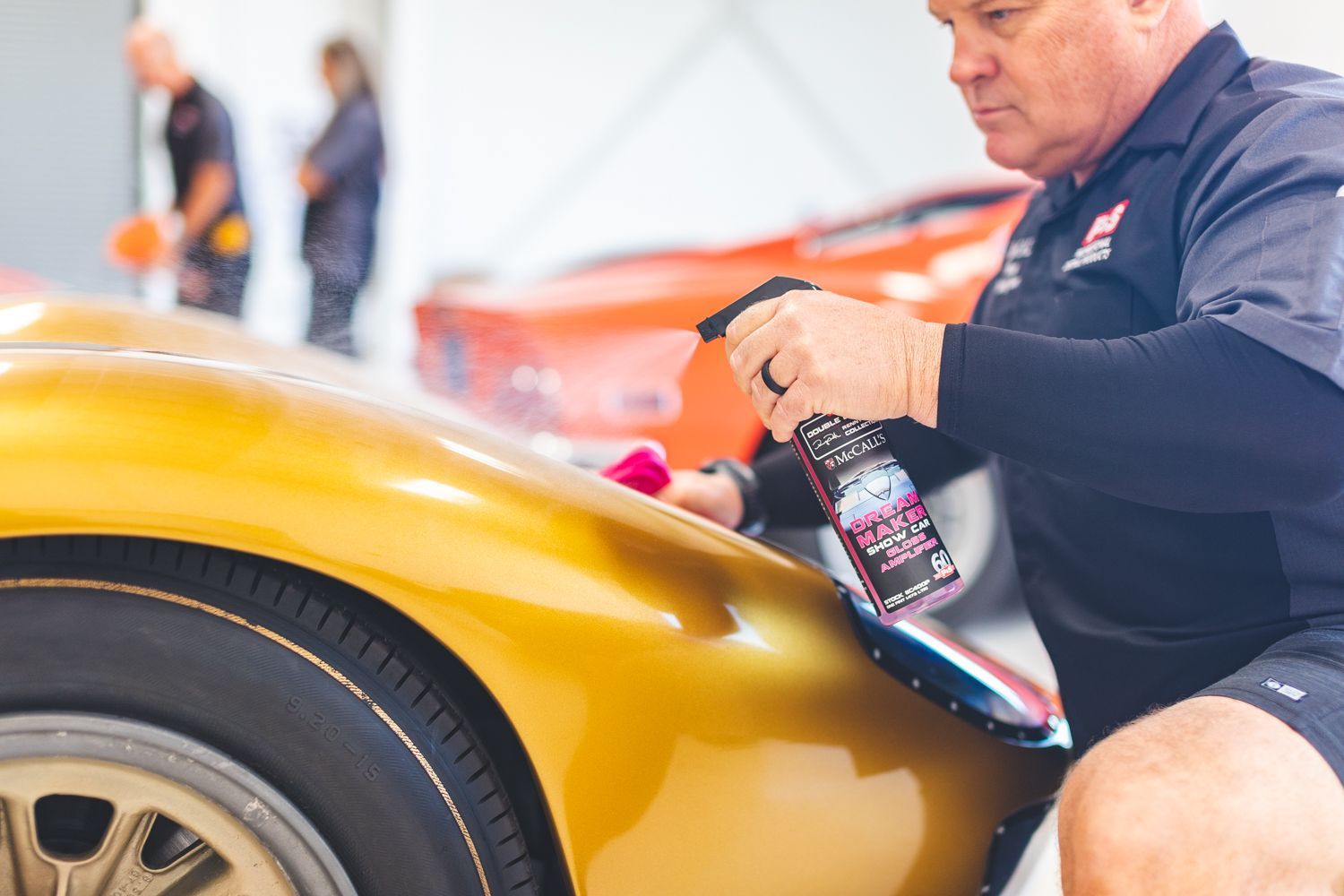 Man spraying cleaner on a gold car in a garage, other people in the background.