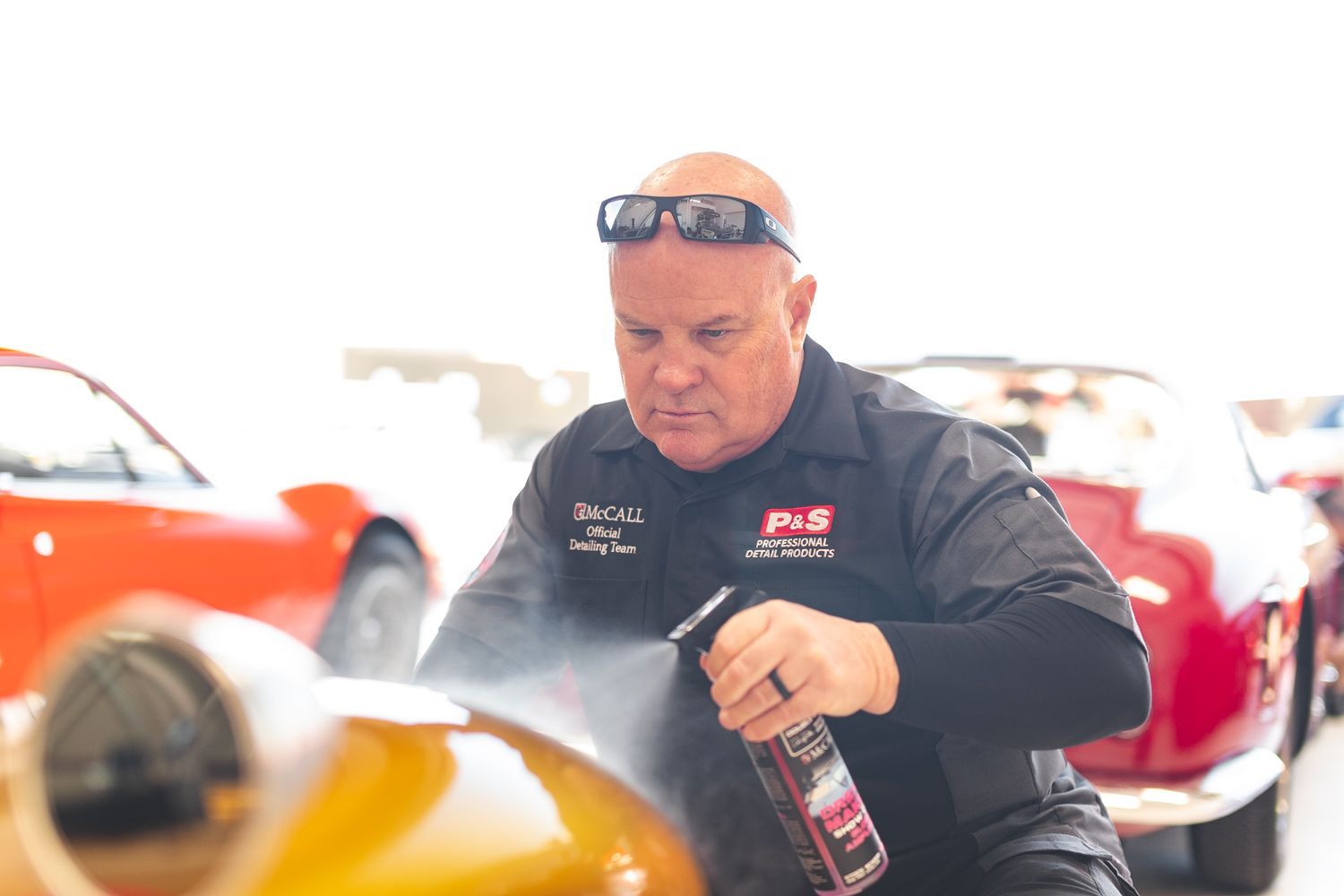 Man spraying a red object, possibly a car. He's outdoors in a garage, red cars in background.
