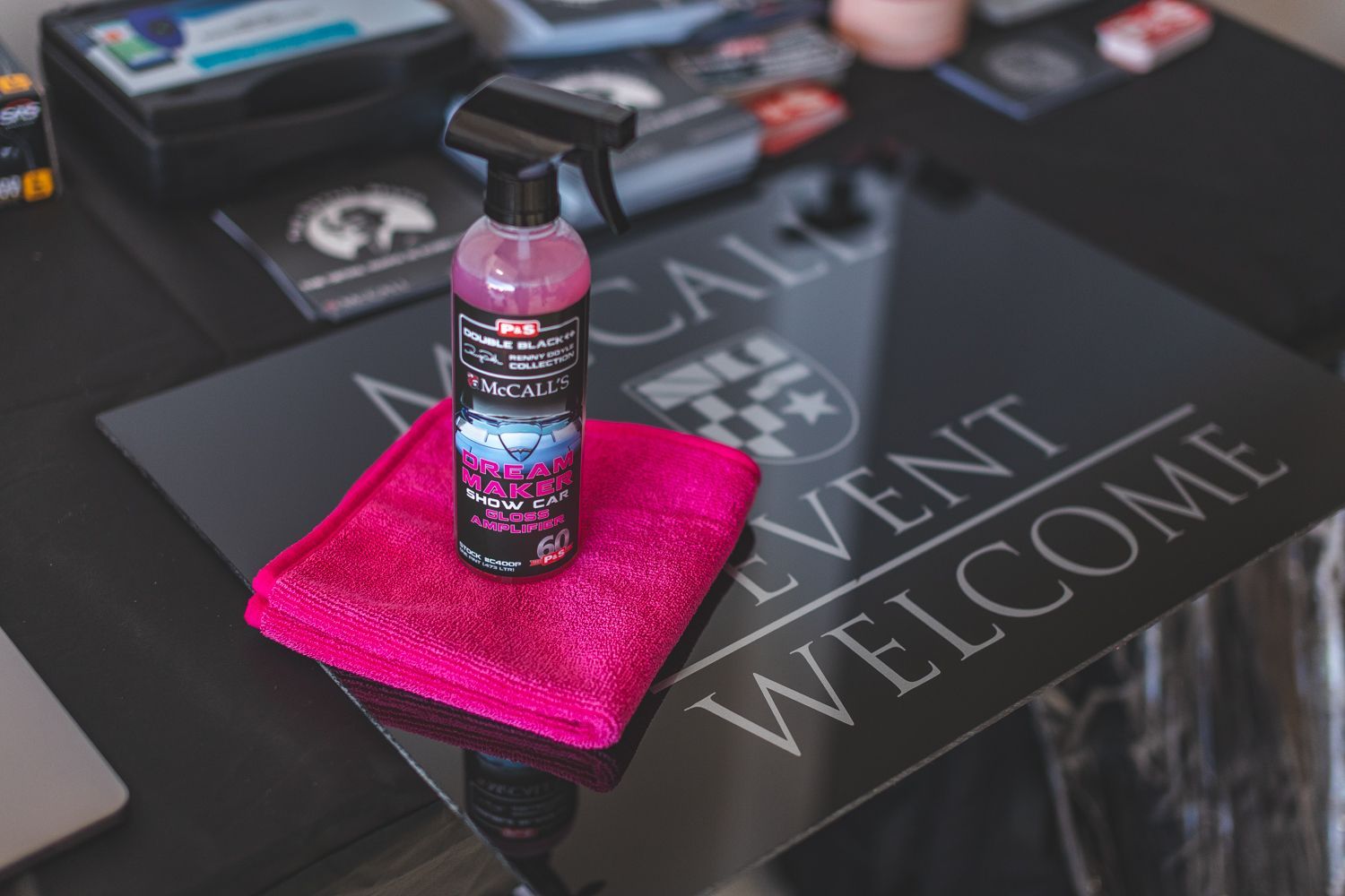 A pink spray bottle, pink cloth, and welcome sign on a black table.