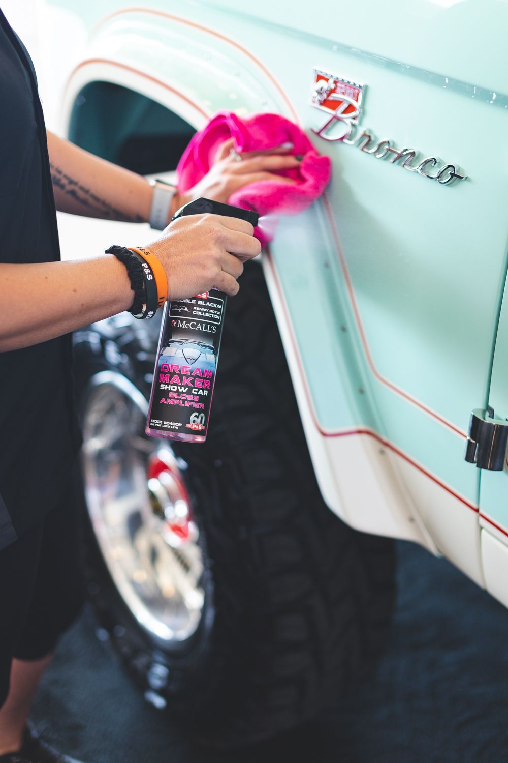 Person cleaning a light blue and white vintage Ford Bronco with a pink microfiber cloth and spray bottle.