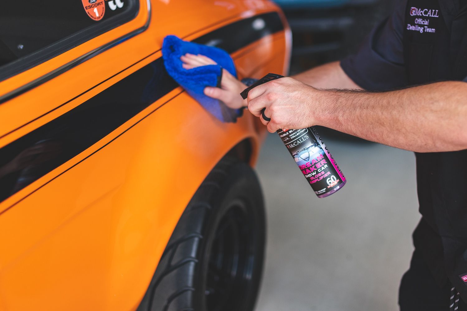Person spraying and wiping orange classic car with black stripes.