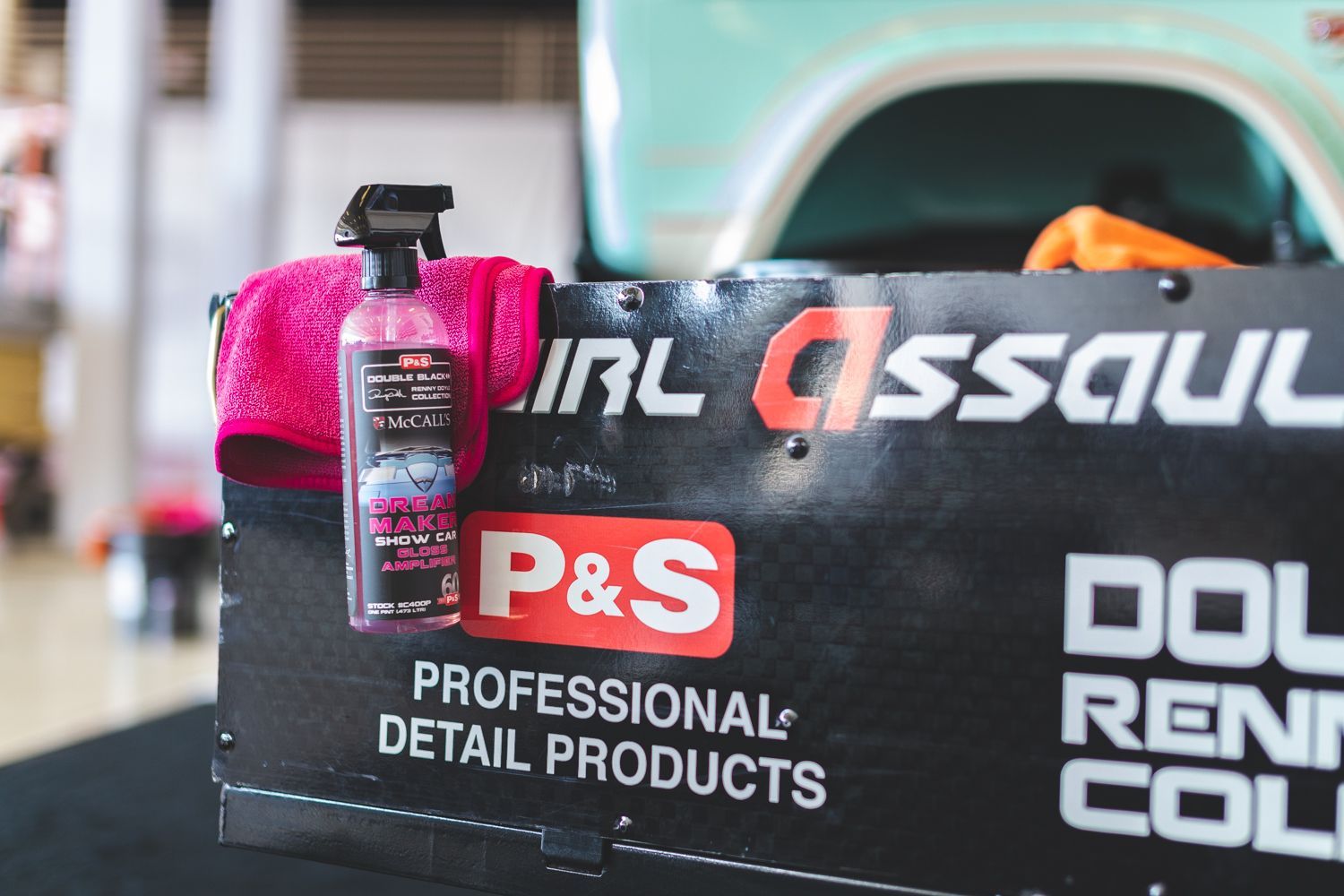 Pink spray bottle and cloth on a black sign for P&S Professional Detail Products with a teal vehicle in the background.