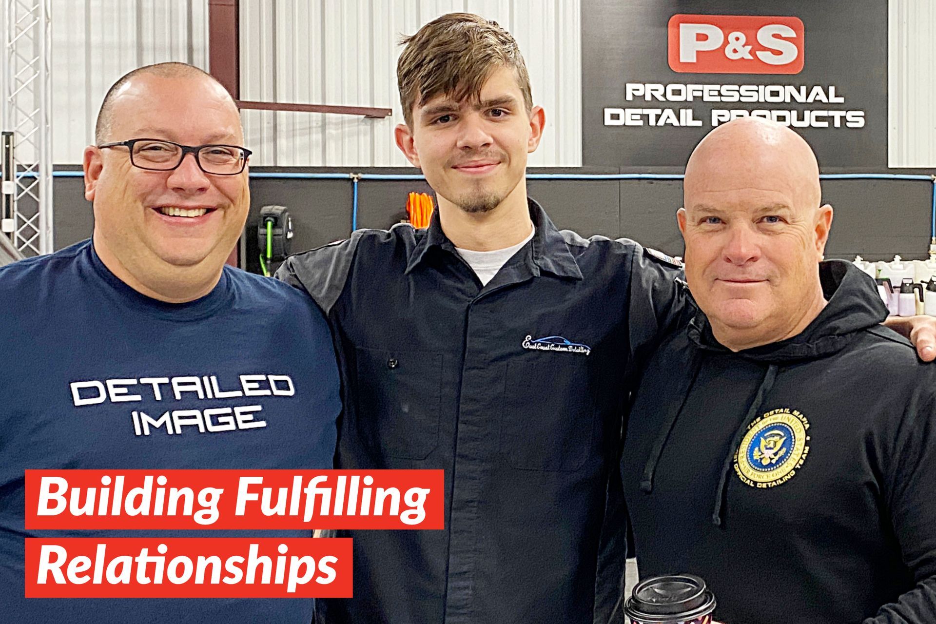 Three men smiling together, inside a warehouse with detailing products in the background. One wears glasses, another a work shirt, and the third a hoodie.
