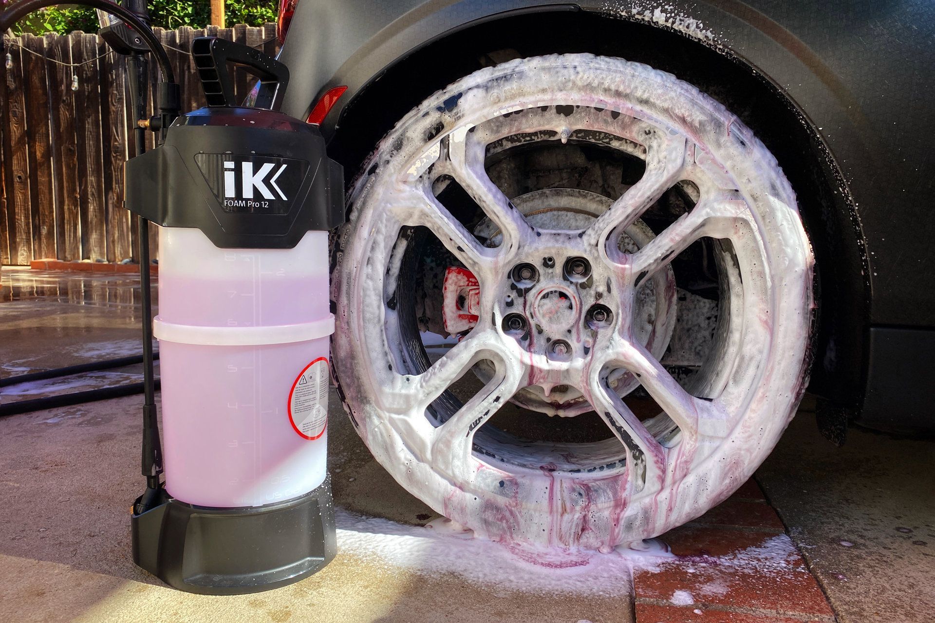 Foamy soap sprayed on a car wheel by a black and white sprayer, outside.