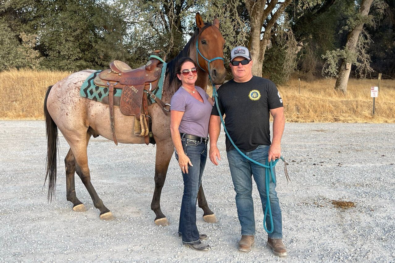 Woman and man pose with a dappled horse wearing a saddle. They stand on gravel outdoors.