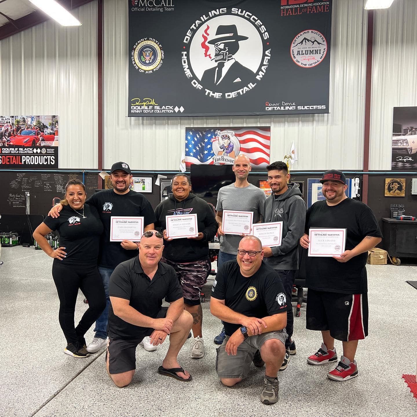 Group of people holding certificates, posing in a garage setting with flags and signage.