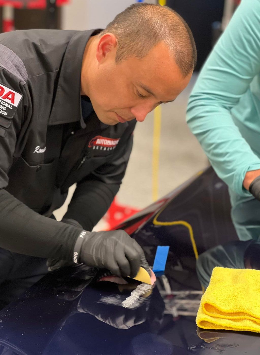 Man in black uniform using a sponge to polish a dark blue car in a garage.