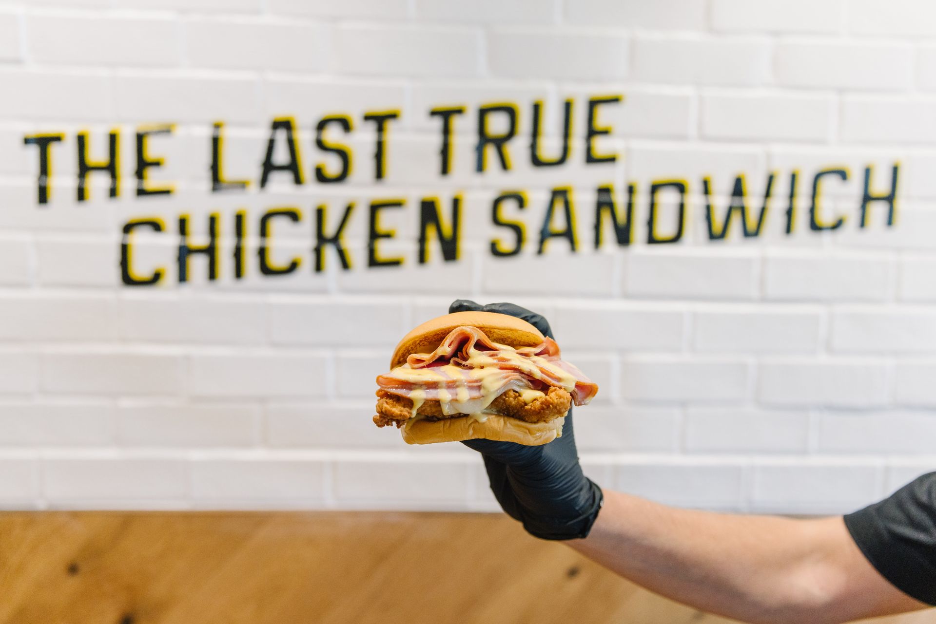 Person holding a fried chicken sandwich with bacon, in front of a sign that reads, 