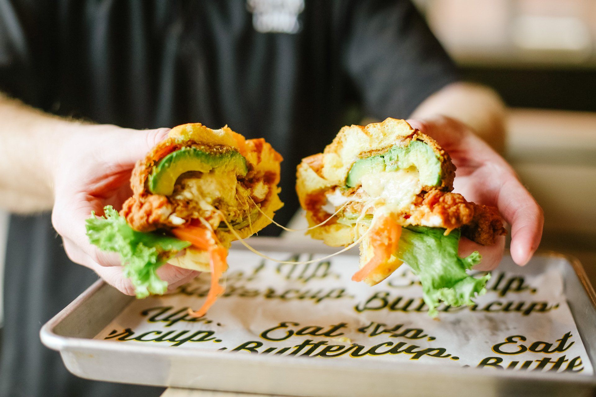 Hands holding a halved fried chicken sandwich with avocado and lettuce, served on a tray.