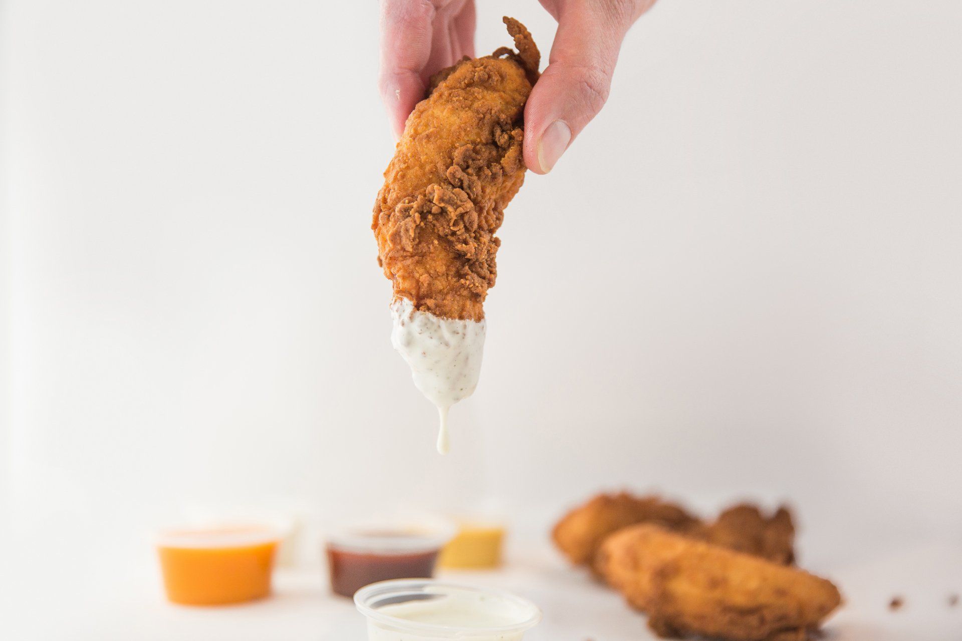 Hand dipping fried chicken tender into white sauce, other sauces and tenders in background.