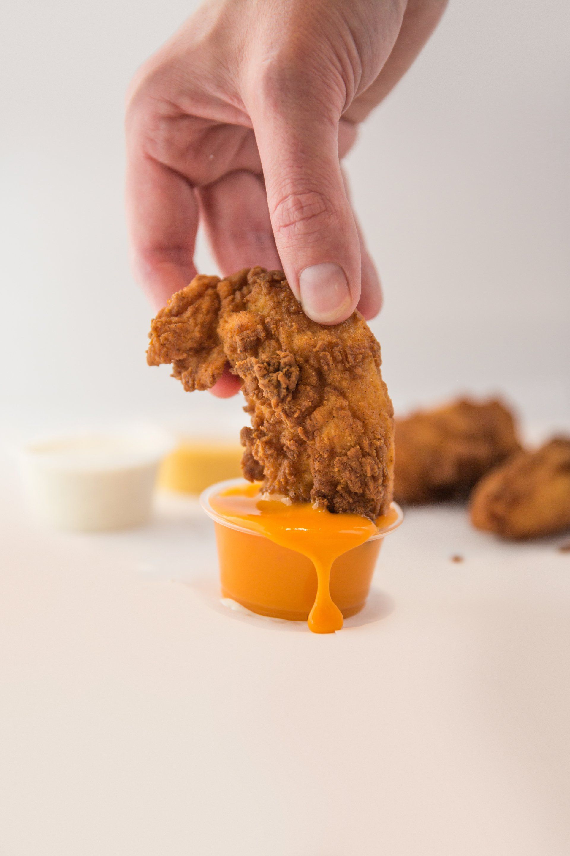 Hand dipping a fried chicken tender into orange sauce. Other tenders and dipping sauces in background.