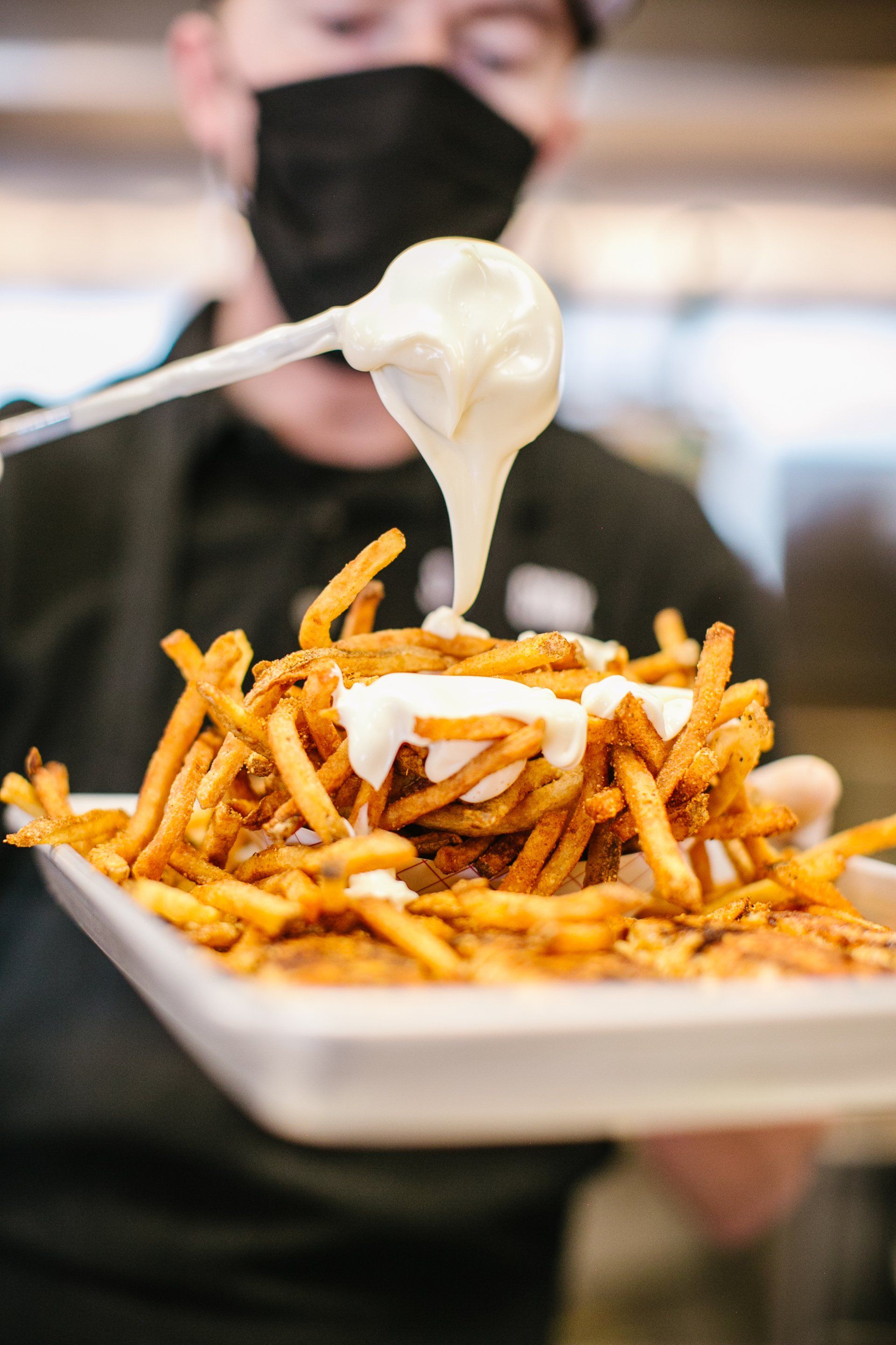 Person in mask pouring creamy sauce over a tray of fries.