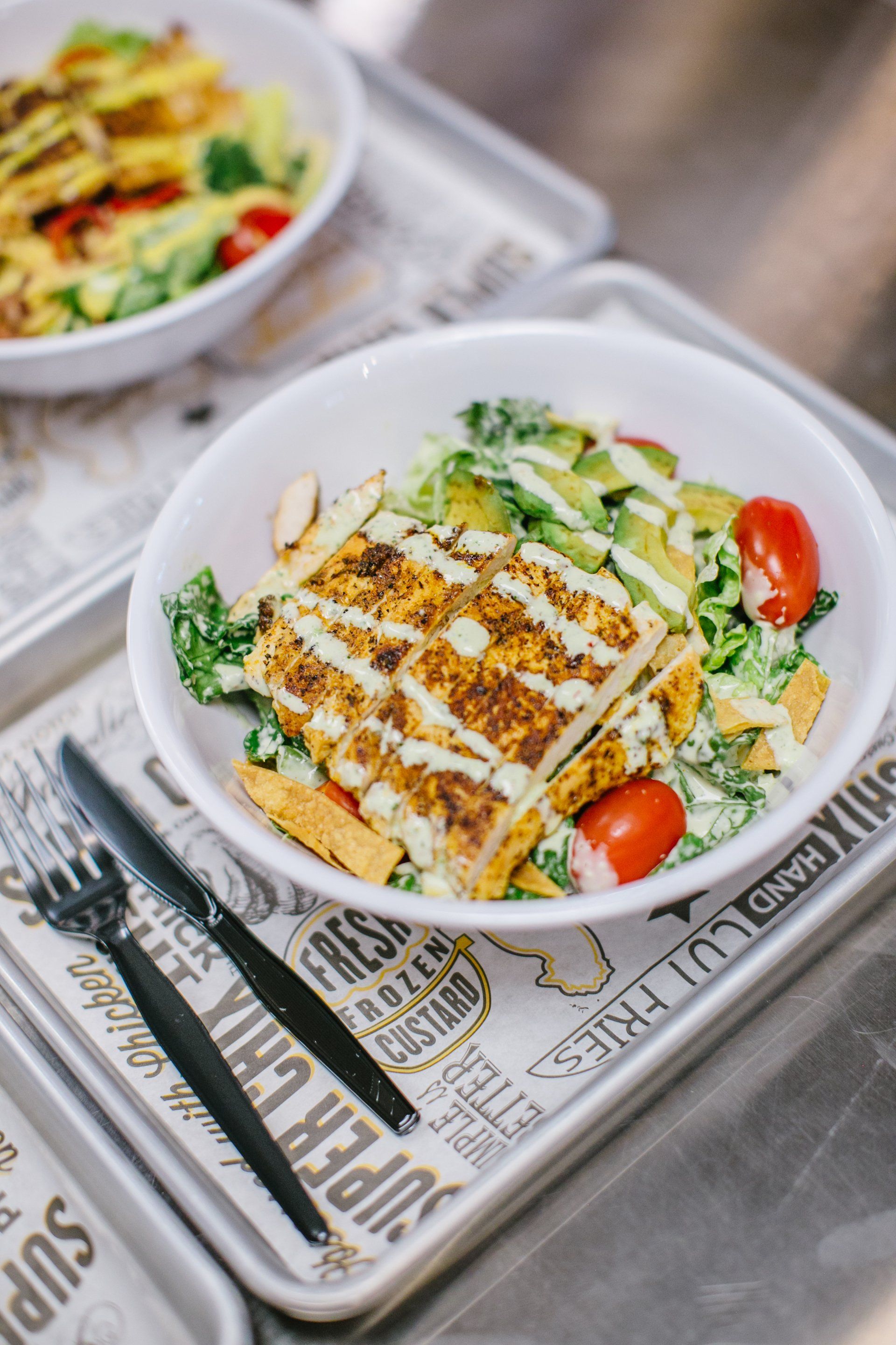 Grilled chicken salad with tomatoes and avocado in a white bowl, on a tray with cutlery.