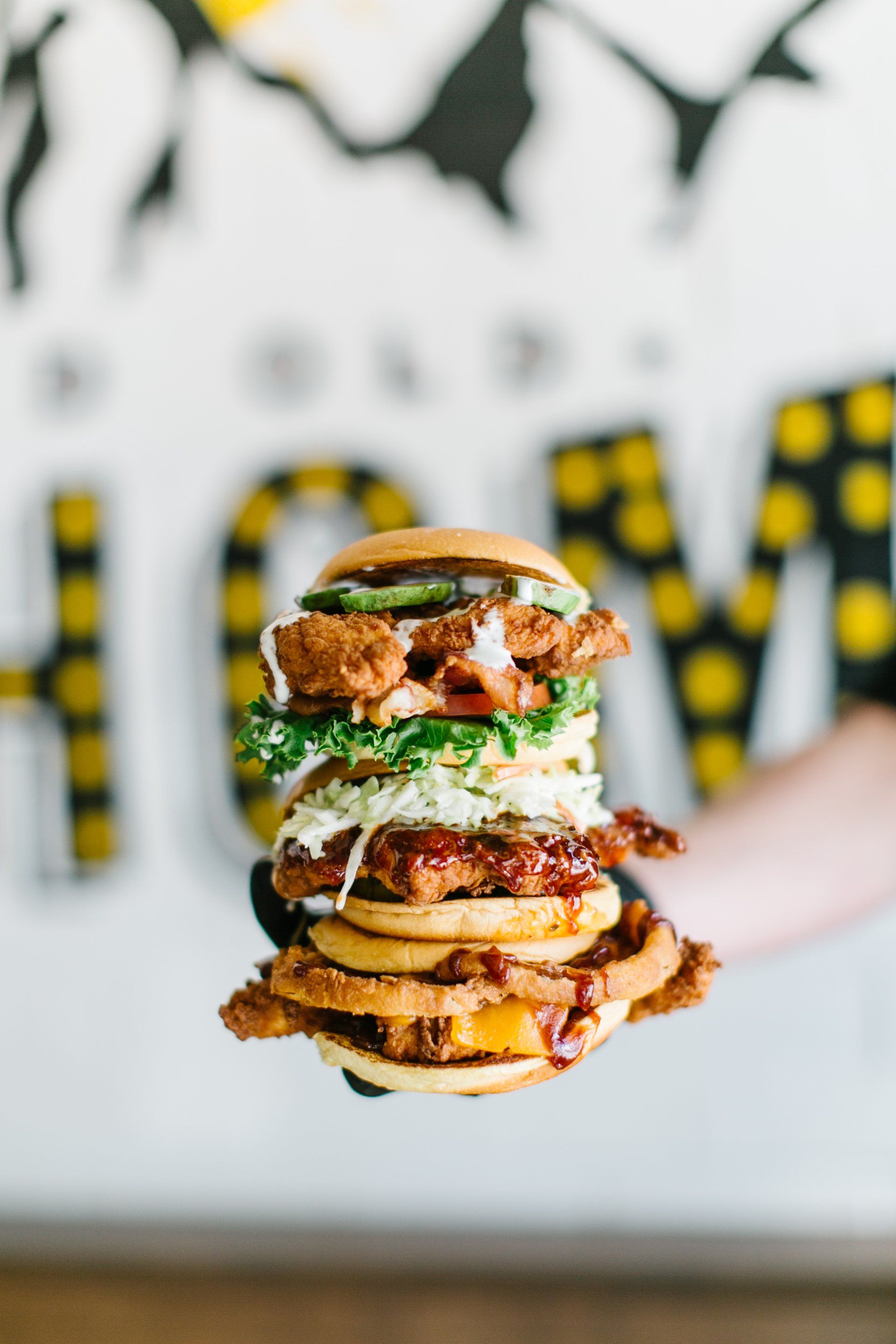 A towering fried chicken sandwich held in black-gloved hands, set against a 