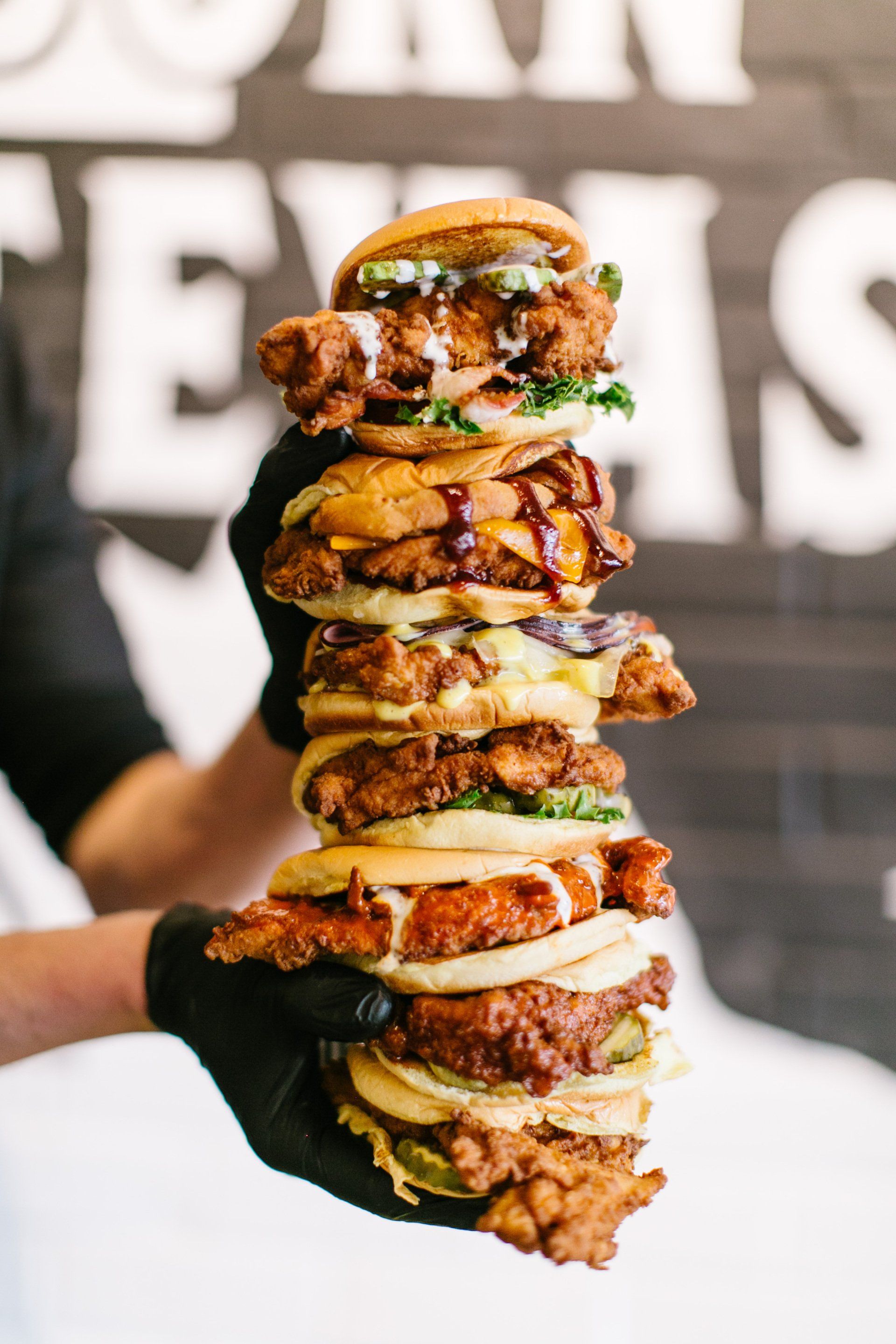 Person in black gloves holding a towering stack of fried chicken sandwiches with various toppings.