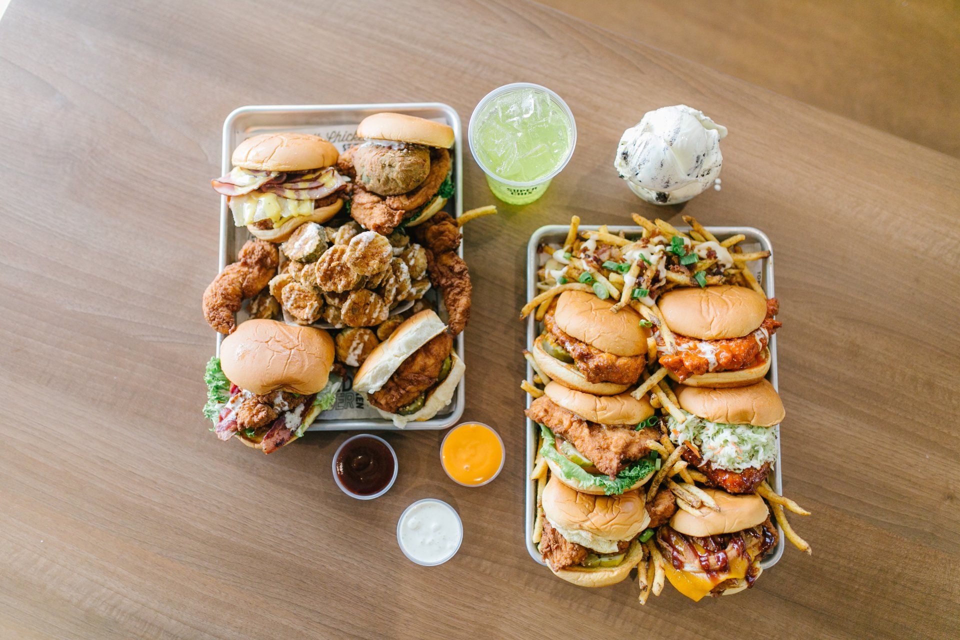 Two trays of burgers, chicken, fries, dips, soda, and ice cream on a wooden table.