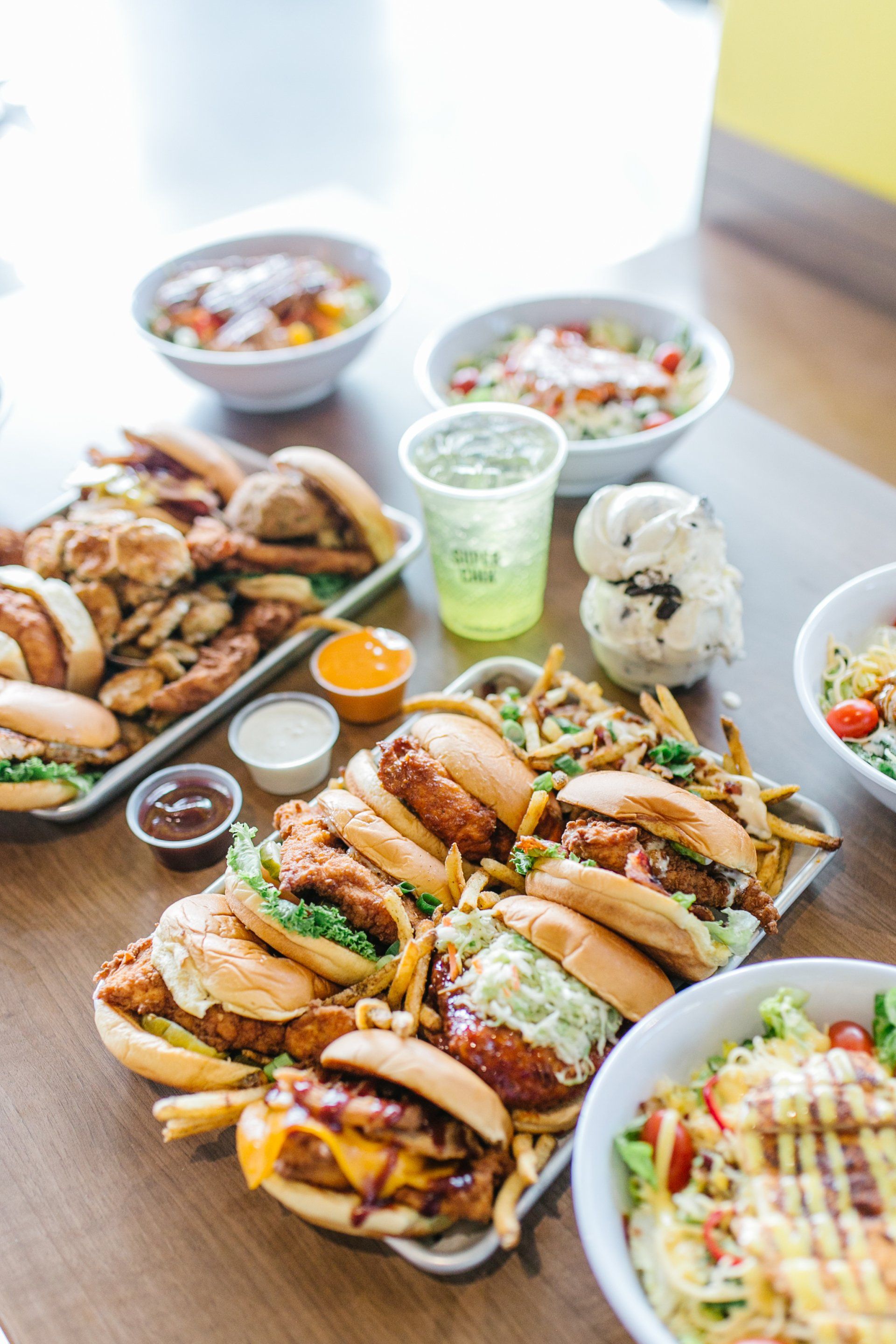 Table of various burgers, salads, fries, and drinks. Food is displayed on trays and bowls.