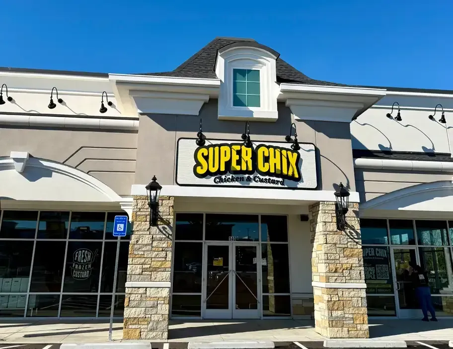 Exterior of Super Chix restaurant with stone columns, sign, and small window. Blue sky.