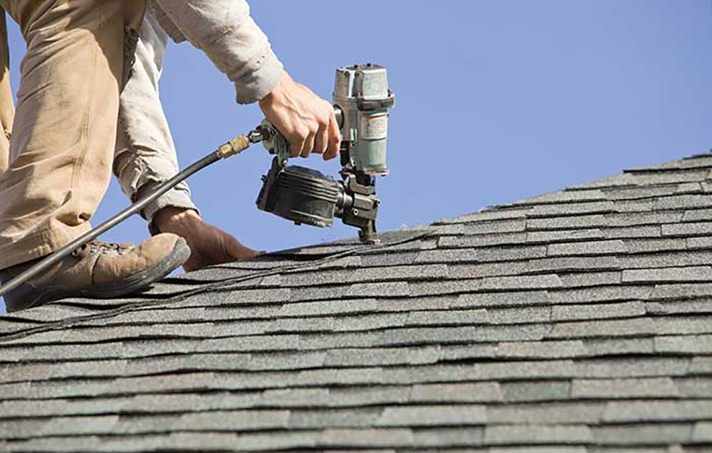 Roofer using a nail gun to install asphalt shingles on a roof against a blue sky.