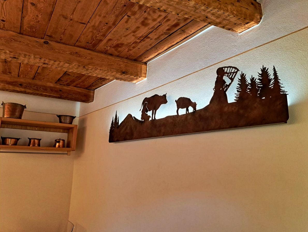 Decorazione murale rustica in metallo raffigurante la silhouette di un paesaggio montuoso con una mucca, una capra e degli alberi, illuminata da dietro.