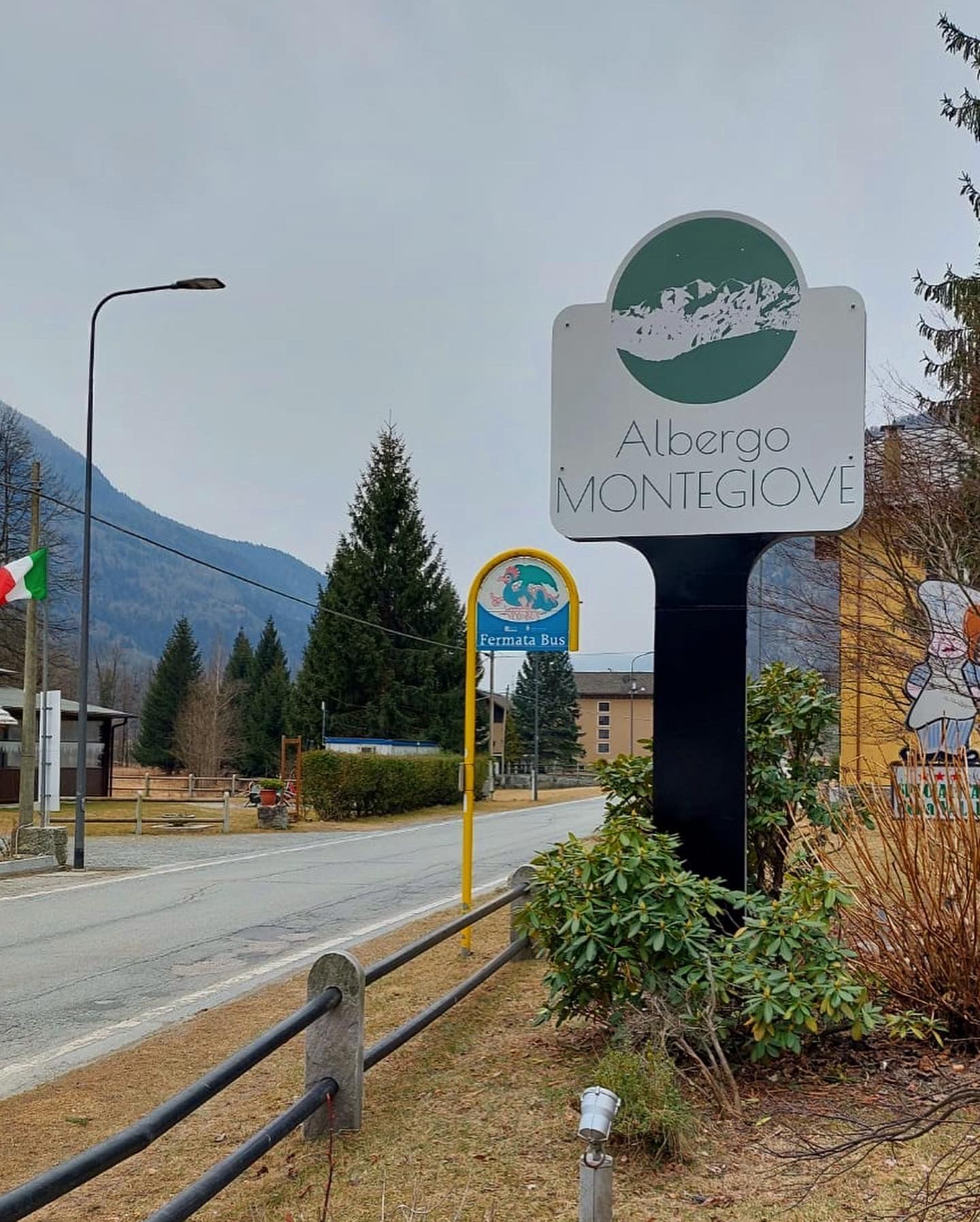 Un cartello bianco con la scritta Albergo Montegiove si erge a lato di una strada con uno sfondo montuoso e un piccolo cartello di fermata dell'autobus.
