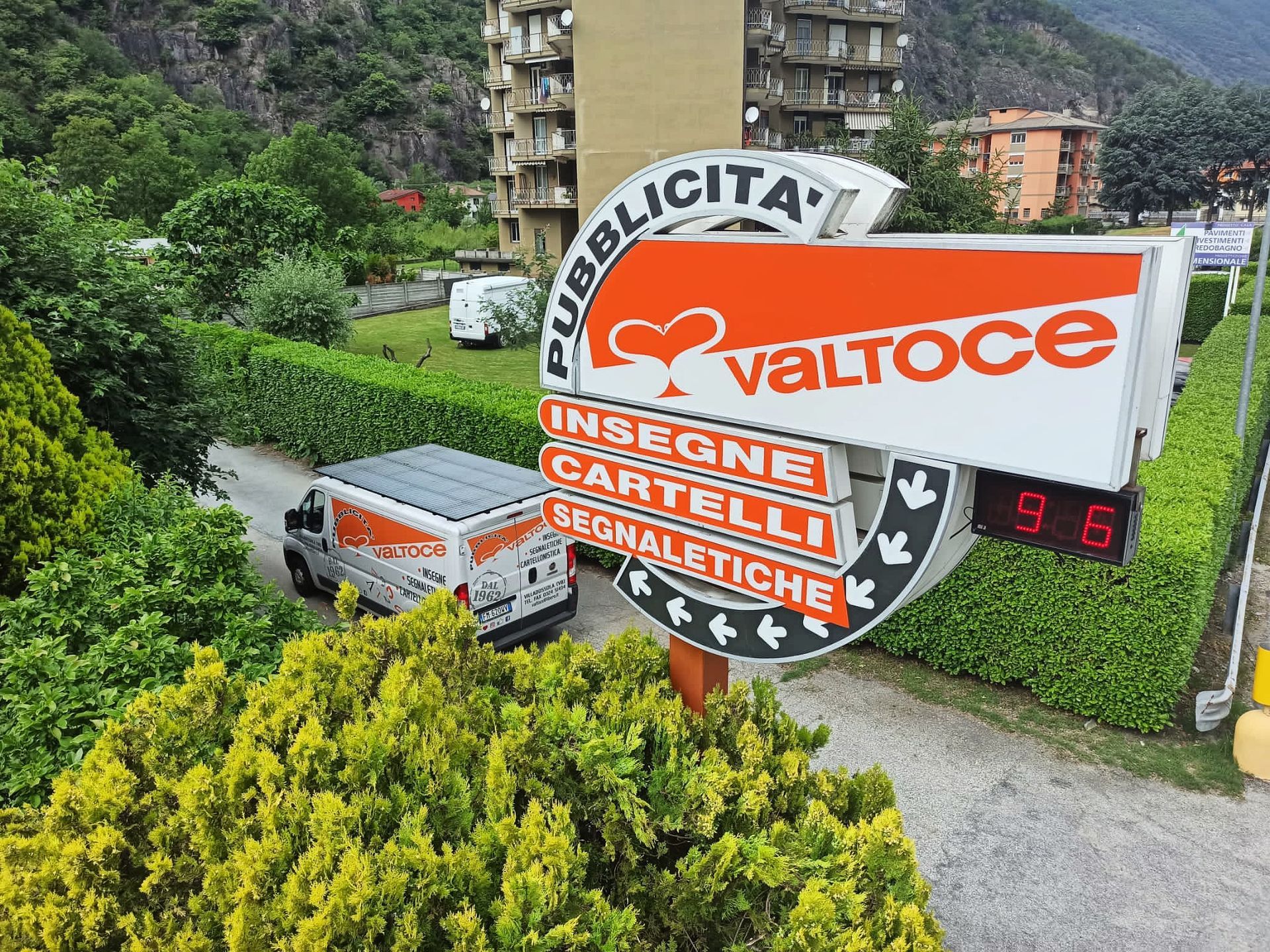 Un cartellone pubblicitario della Valtoce con un furgone bianco che passa su una strada incorniciata da siepi verdi ed edifici sullo sfondo.