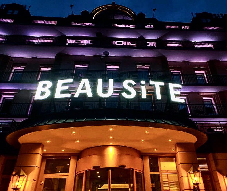 Una veduta notturna dal basso dell'ingresso illuminato dell'hotel BEAUSITE, caratterizzato da lettere bianche luminose e calde luci d'accento.