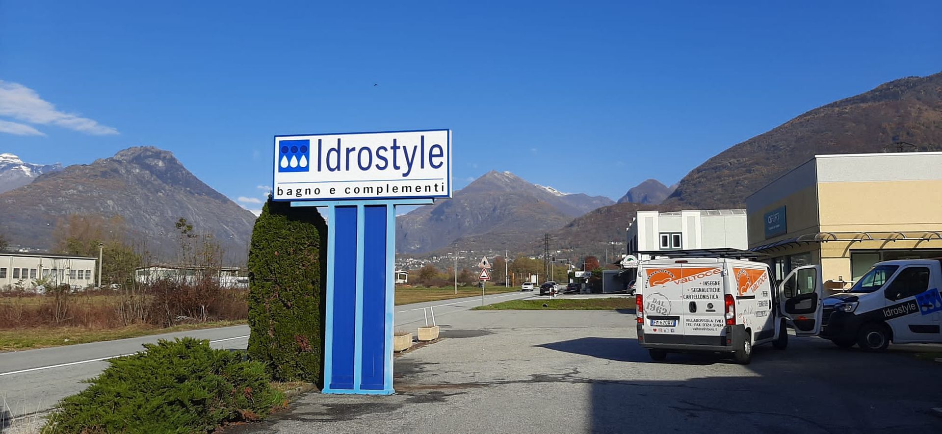 Un'insegna dell'attività commerciale Idrastyle si erge in un parcheggio sterrato, con furgoni parcheggiati accanto a un edificio sullo sfondo di montagne.