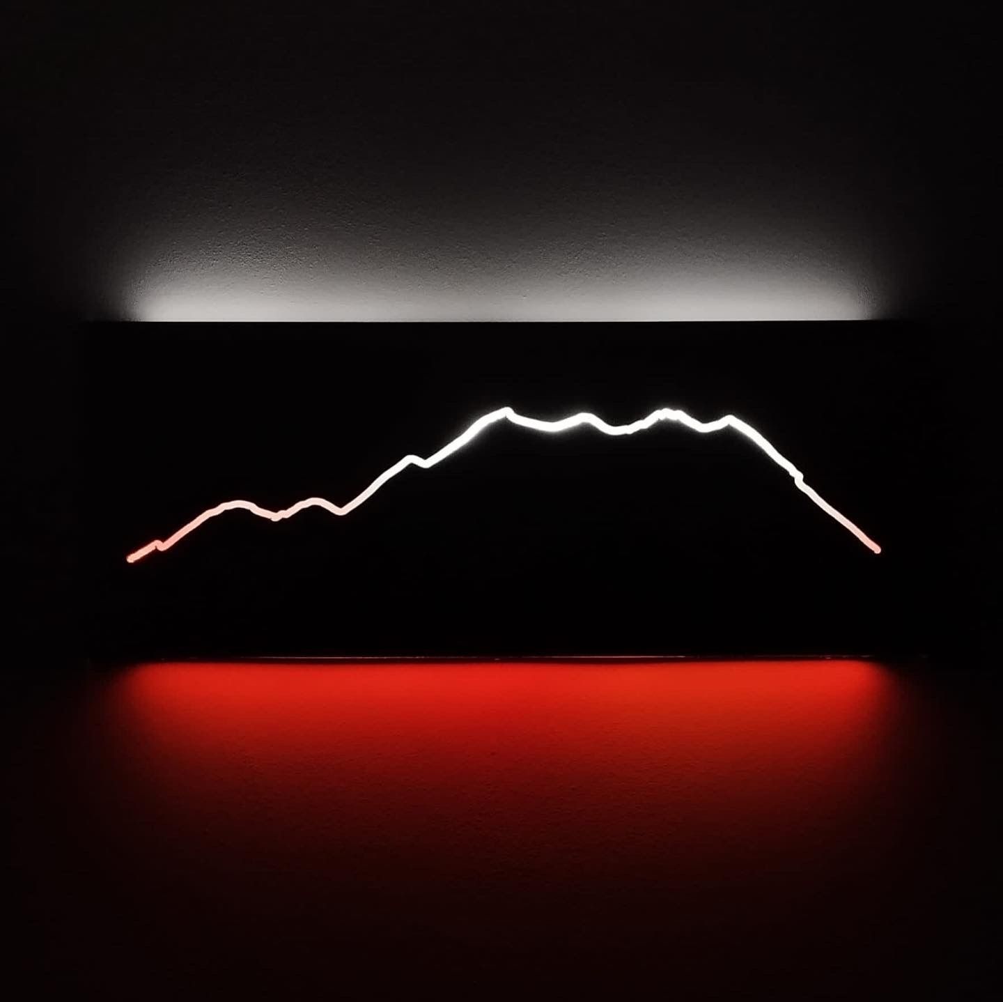 Un'insegna a muro scura con una linea bianca illuminata che rappresenta la sagoma di una montagna, che brilla sopra una striscia luminosa rossa.
