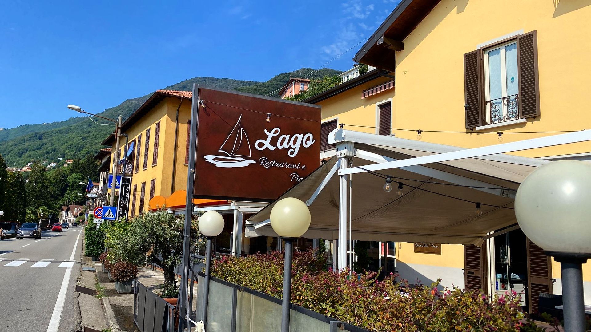 In una giornata di sole, un ristorante di colore giallo chiamato Lago, con un patio esterno, un'insegna ben visibile e le montagne sullo sfondo.