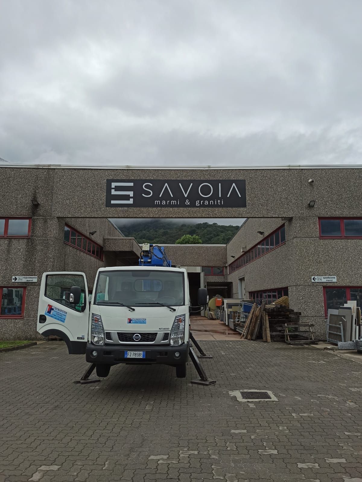 Un camioncino bianco con piattaforma aerea è parcheggiato davanti a un edificio industriale in pietra con la scritta 