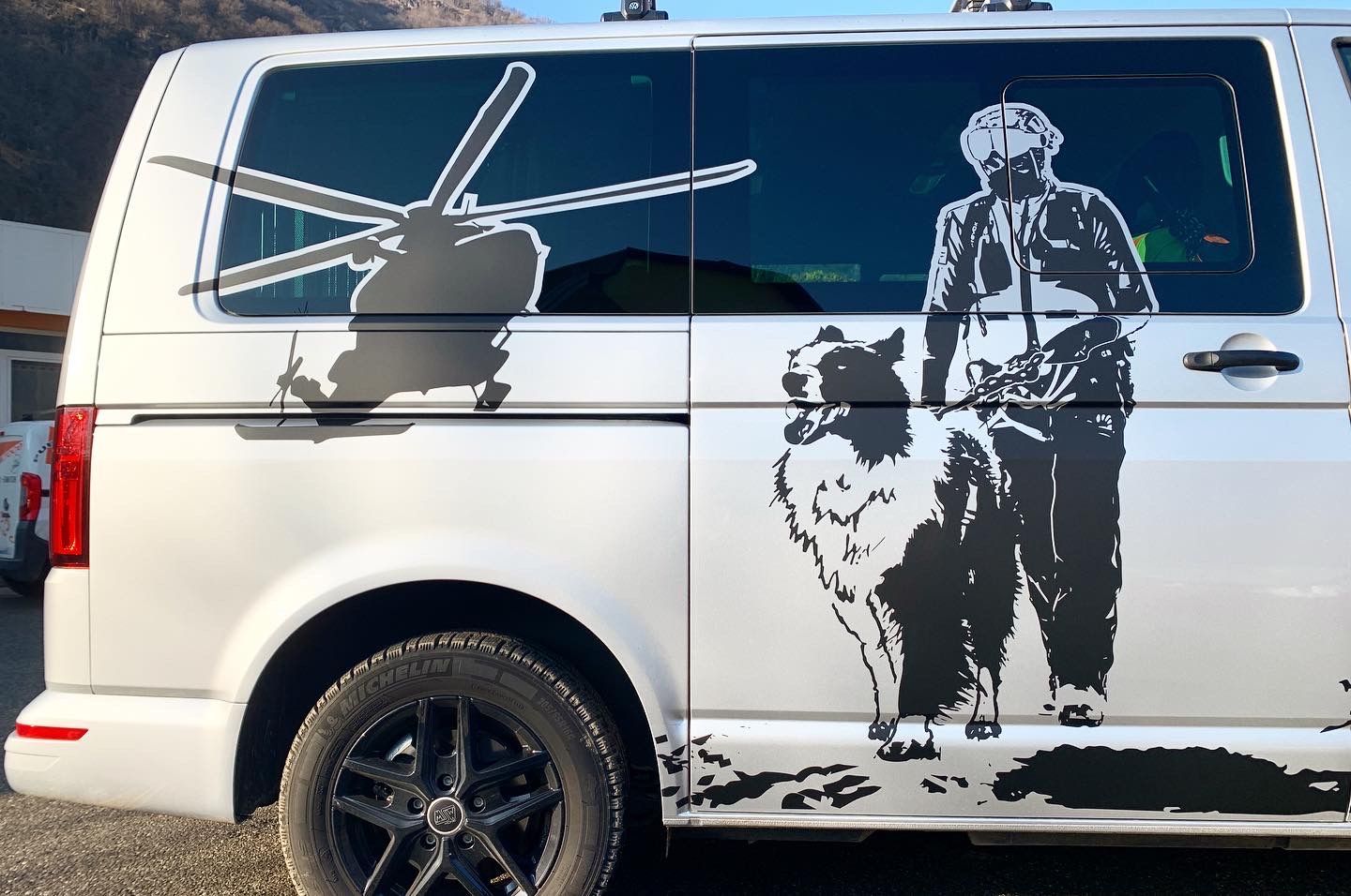 Un furgone argentato presenta adesivi in ​​vinile nero raffiguranti un elicottero e un addestratore con un cane, in stile silhouette.
