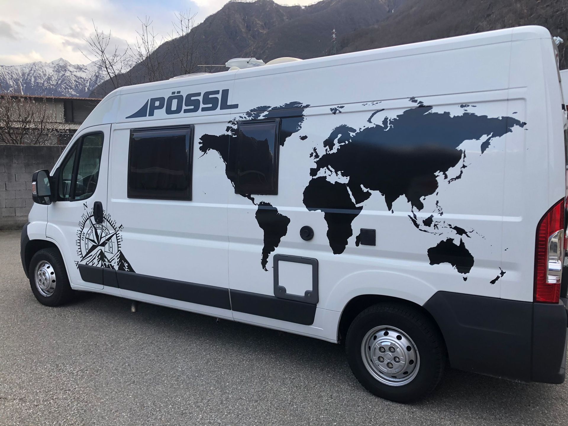 Un camper Pössl bianco con un adesivo nero raffigurante una mappa del mondo e una bussola, parcheggiato sullo sfondo di una montagna.