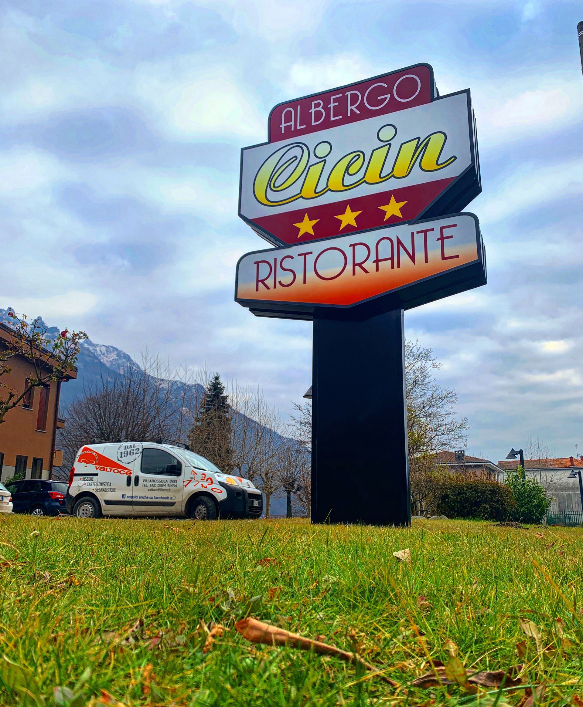 Un'insegna con tre stelle per l'Albergo Cicin Ristorante si erge in un prato, con le montagne sullo sfondo e un furgone bianco per le consegne.