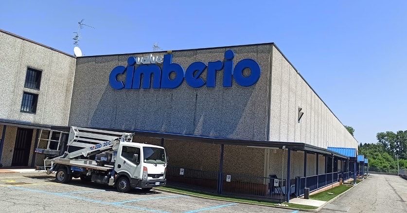 Un camioncino bianco con gru è parcheggiato davanti a un edificio industriale beige con il logo blu 