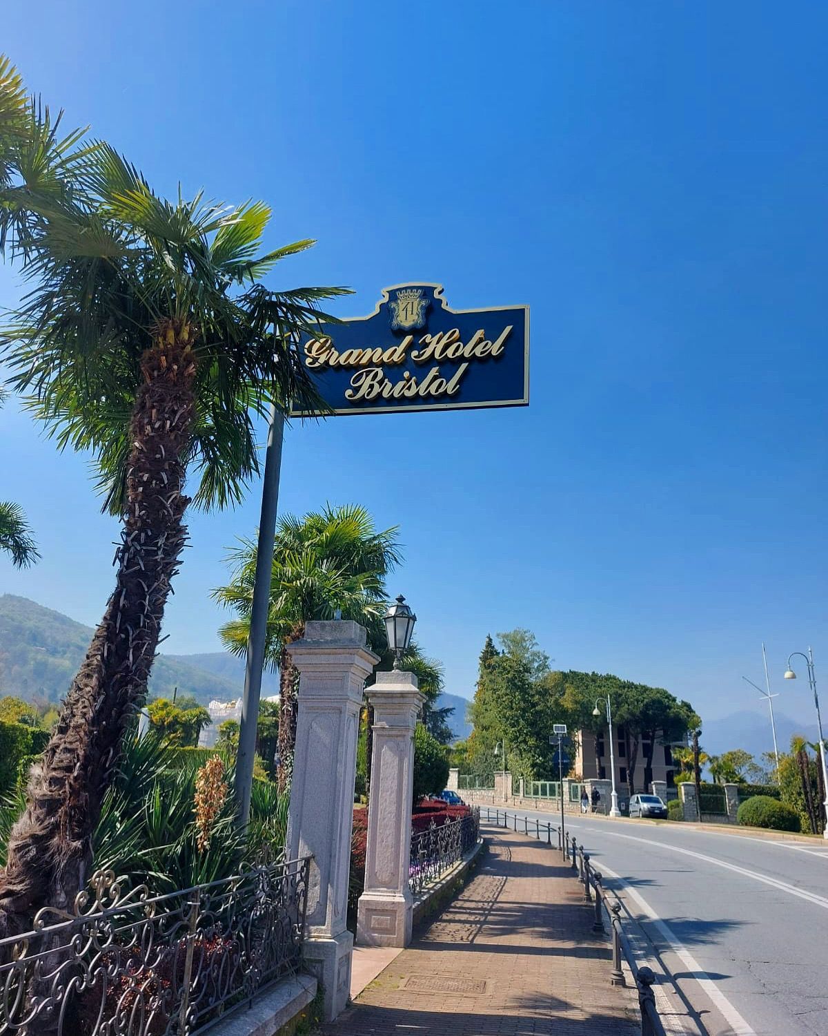 Un'insegna del Grand Hotel Bristol si erge su un marciapiede fiancheggiato da palme lungo una strada soleggiata.