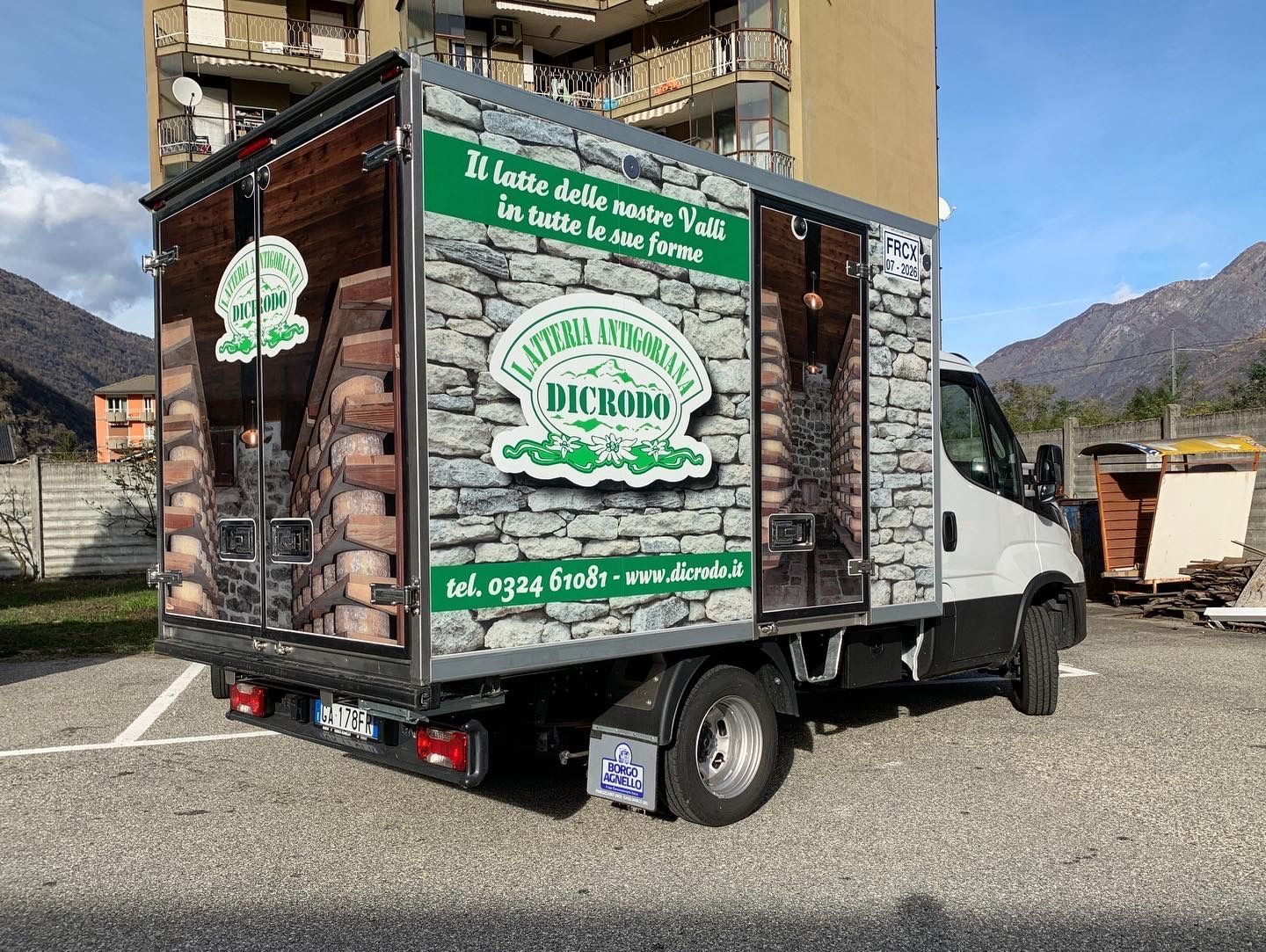 Un furgone bianco parcheggiato all'aperto, con stampe raffiguranti forme di formaggio impilate e muri in pietra, tipico di un'azienda casearia.
