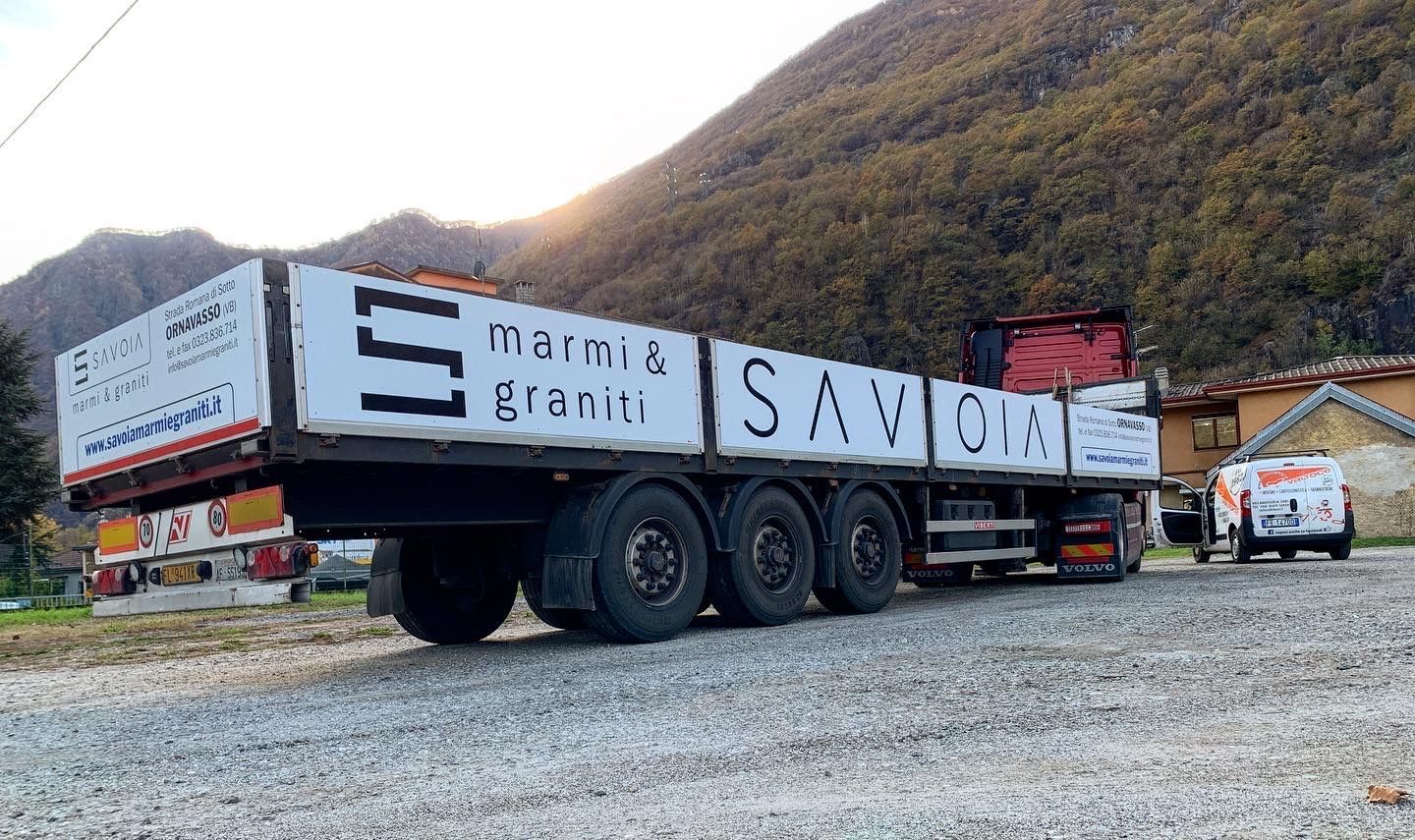 Un grande camion bianco a pianale ribassato, parcheggiato su ghiaia in una zona montuosa, recava la scritta 